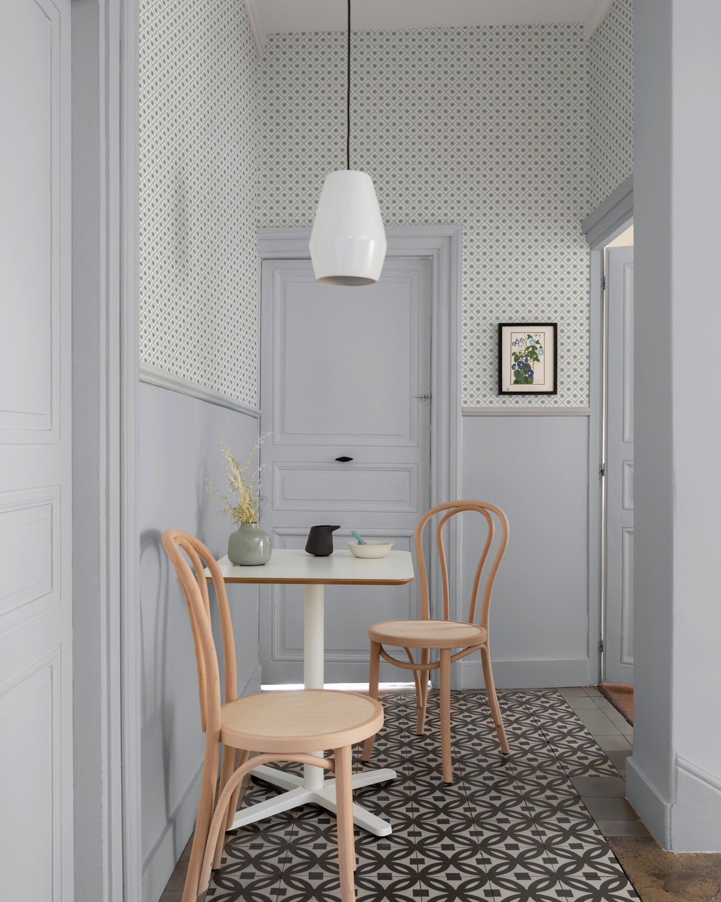 Coin repas aux murs gris perle et papier peint Maître Domino Libéron, avec table blanche, chaises en bois et suspension blanche.