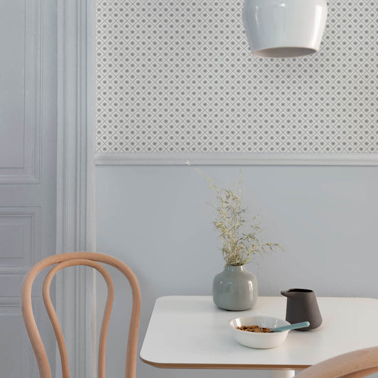 Coin repas aux murs bleu gris et motif géométrique Maître Domino Libéron, avec table blanche, chaises en bois clair et vase.