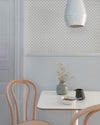 Coin repas aux murs bleu gris et motif géométrique Maître Domino Libéron, avec table blanche, chaises en bois clair et vase.