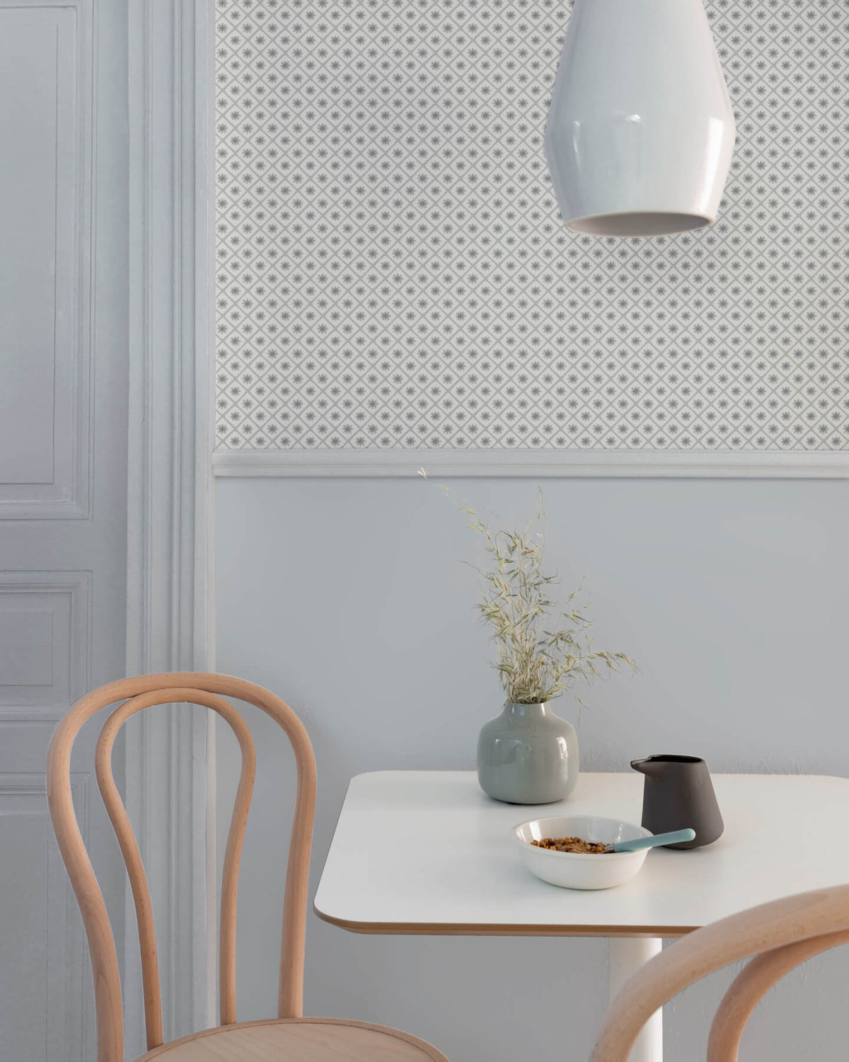 Coin repas aux murs bleu gris et motif géométrique Maître Domino Libéron, avec table blanche, chaises en bois clair et vase.