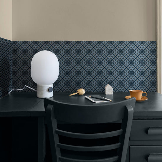 Coin bureau aux murs beige et motif géométrique bleu Maître Domino Libéron, avec chaise sombre, lampe blanche et tasse.