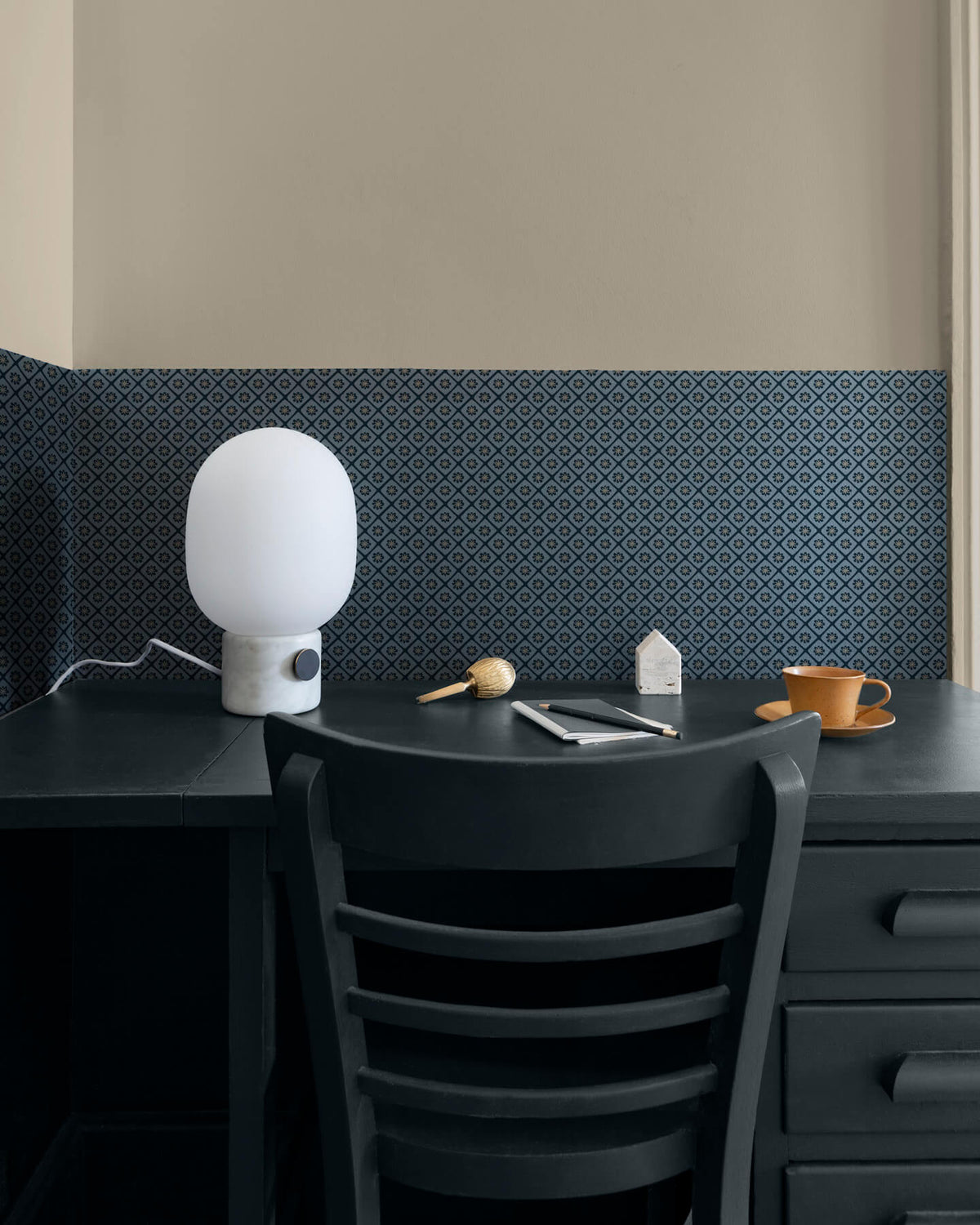 Coin bureau aux murs beige et motif géométrique bleu Maître Domino Libéron, avec chaise sombre, lampe blanche et tasse.