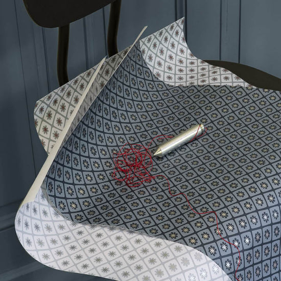 Papier peint Maître Domino Libéron aux motifs bleu grisé, déployé sur une chaise noire avec stylo et fil rouge.