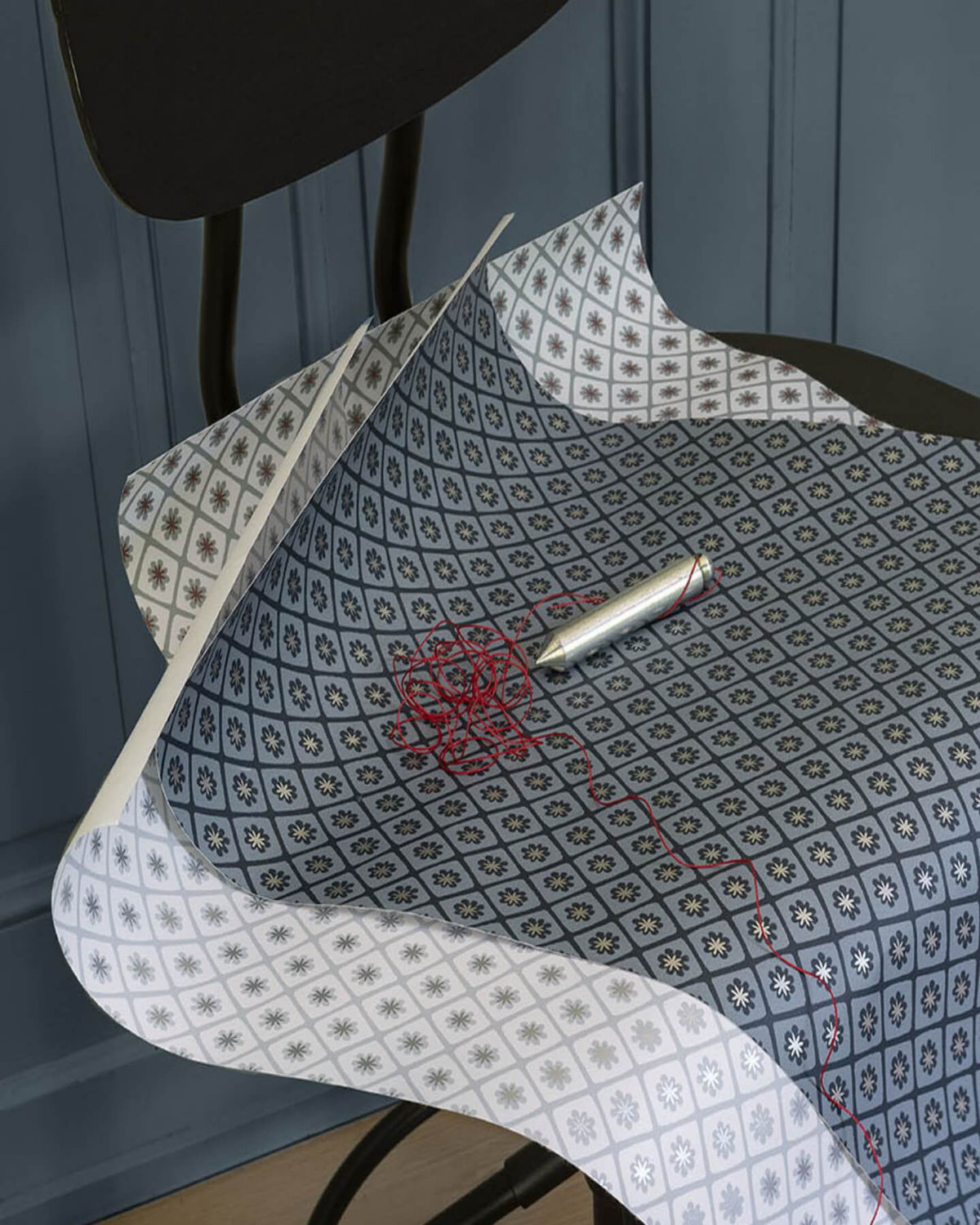 Papier peint Maître Domino Libéron aux motifs bleu grisé, déployé sur une chaise noire avec stylo et fil rouge.