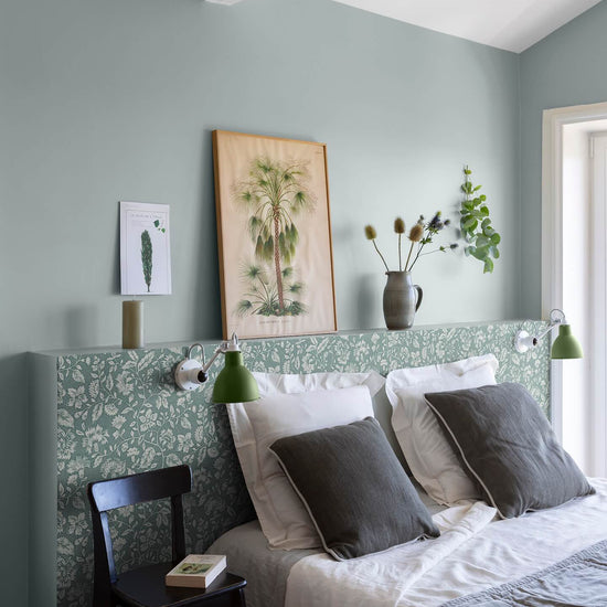 Chambre aux murs vert d’eau et tête de lit florale, teinte Œillet Reine Libéron, avec linge blanc, chaise noire et lampes vertes.