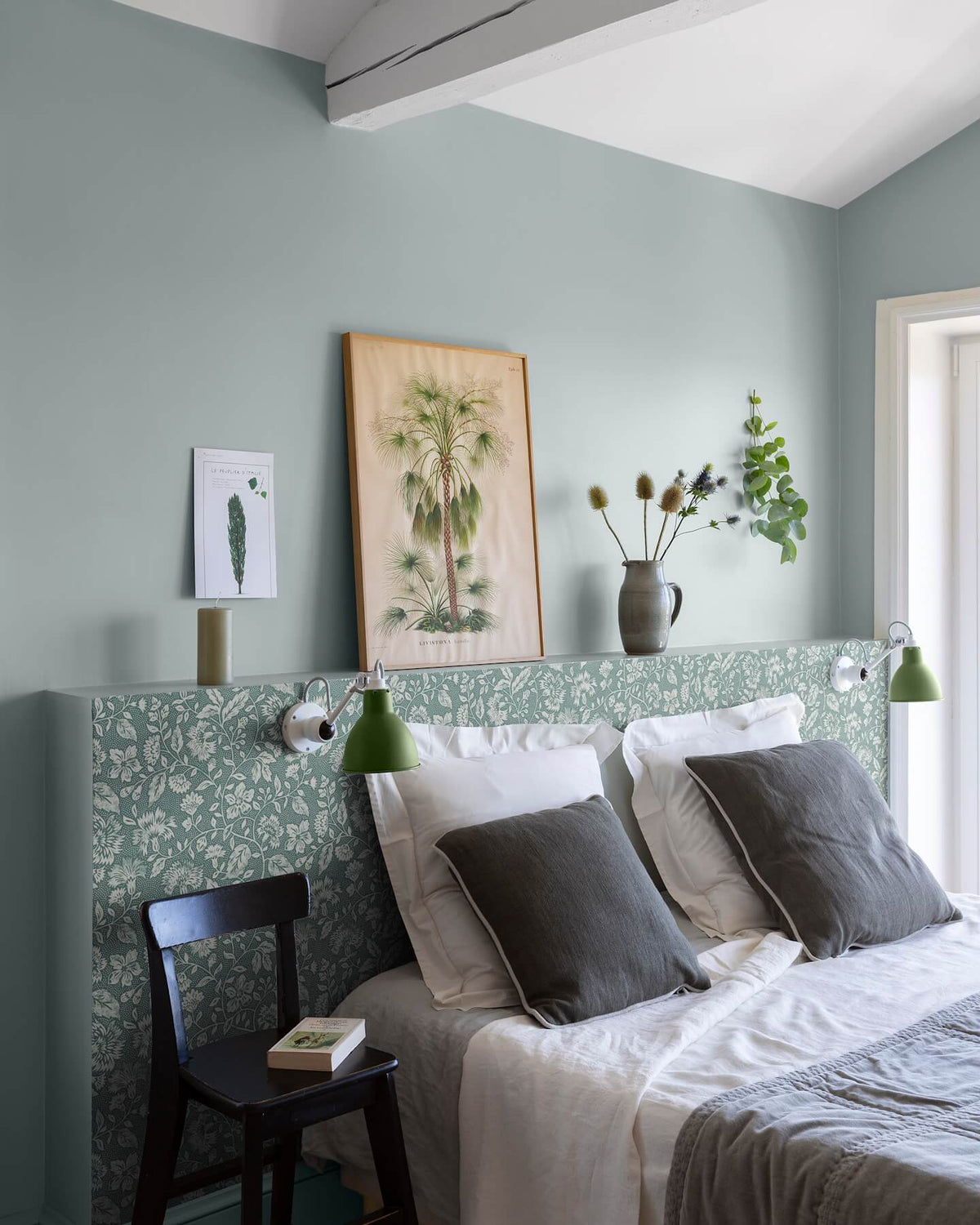 Chambre aux murs vert d’eau et tête de lit florale, teinte Œillet Reine Libéron, avec linge blanc, chaise noire et lampes vertes.
