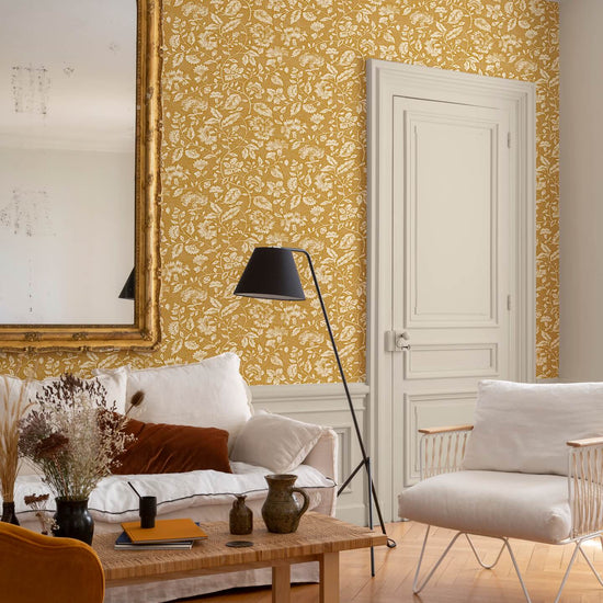 Salon au mur floral ocre et blanc, teinte Œillet Reine Libéron, avec miroir doré, canapé clair et fauteuil tressé.