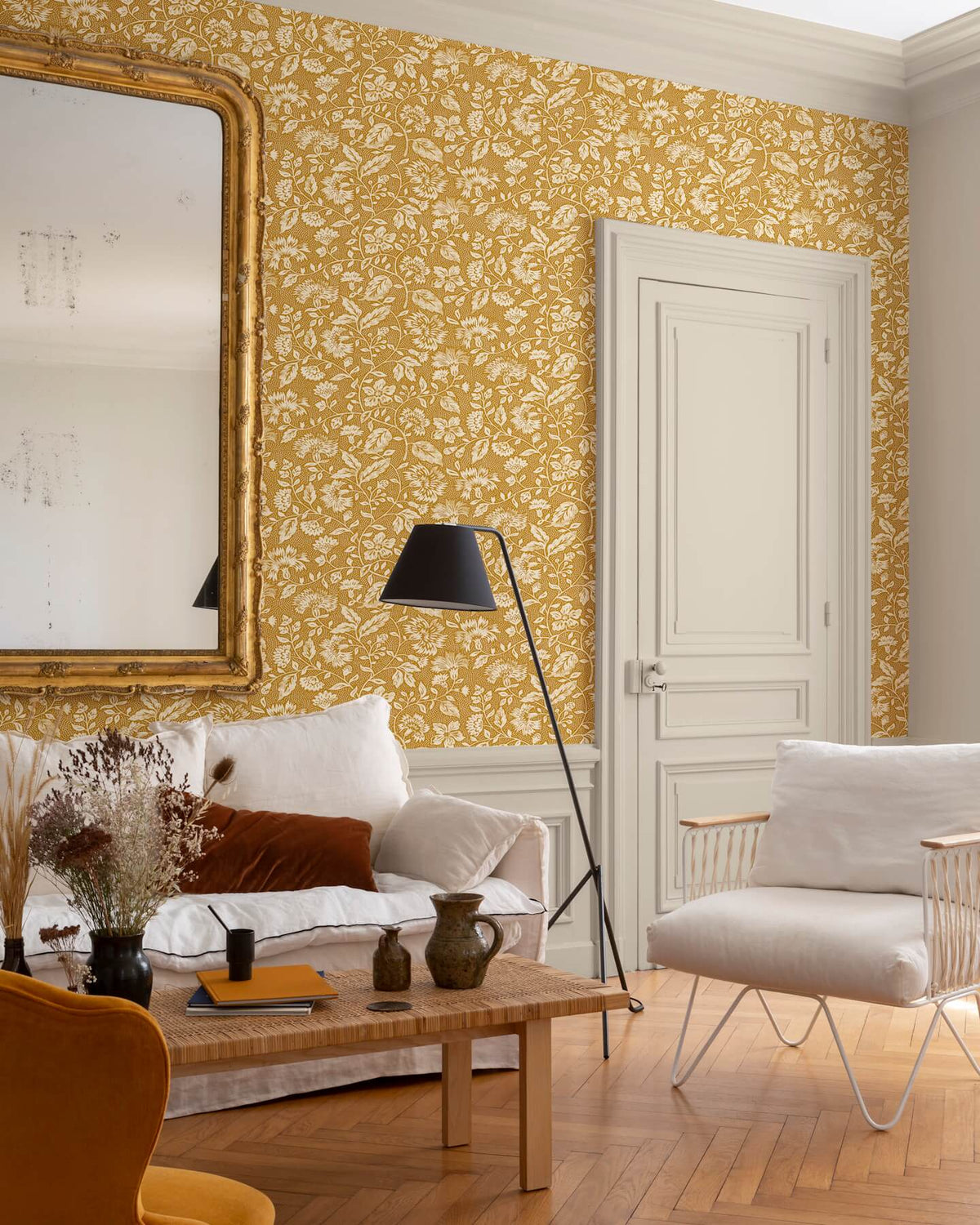 Salon au mur floral ocre et blanc, teinte Œillet Reine Libéron, avec miroir doré, canapé clair et fauteuil tressé.