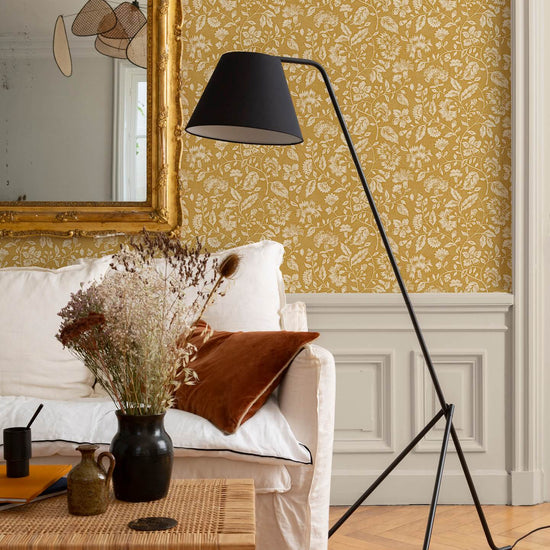 Détail de salon au mur floral ocre et blanc, teinte Œillet Reine Libéron, avec miroir doré, canapé clair et lampe noire.