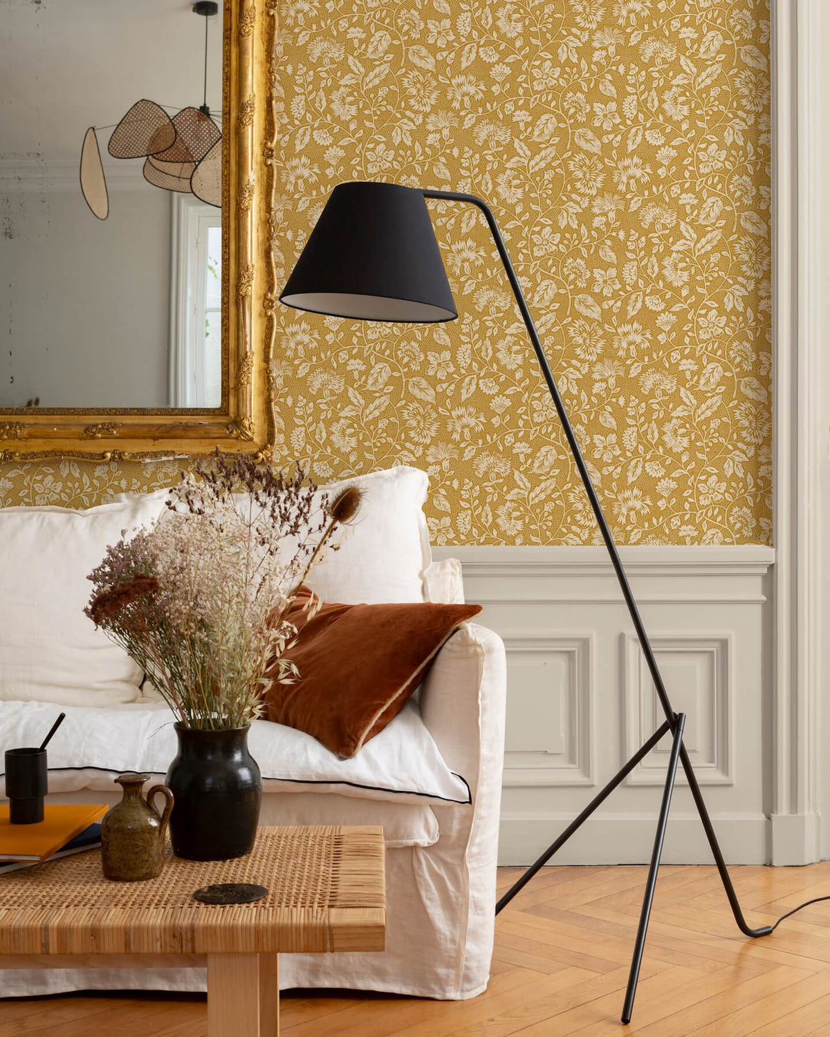 Détail de salon au mur floral ocre et blanc, teinte Œillet Reine Libéron, avec miroir doré, canapé clair et lampe noire.