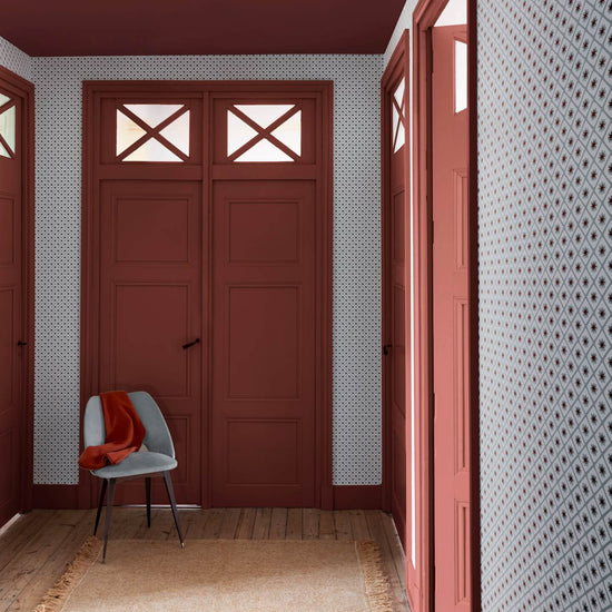 Entrée aux murs terracotta et papier peint Maître Domino Libéron, avec portes assorties, chaise grise et tapis beige.