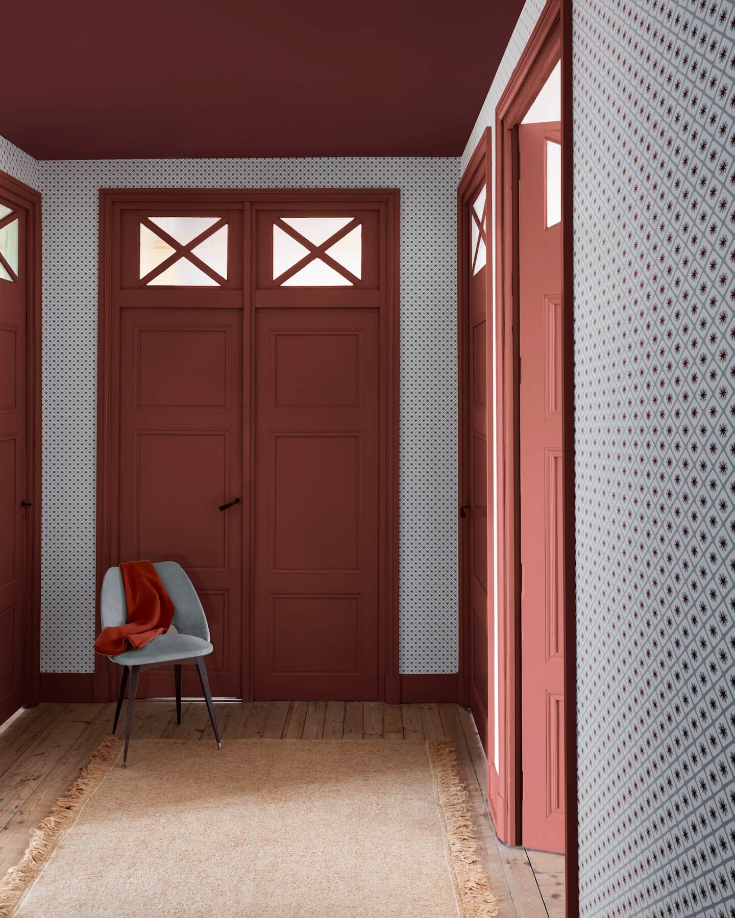 Entrée aux murs terracotta et papier peint Maître Domino Libéron, avec portes assorties, chaise grise et tapis beige.