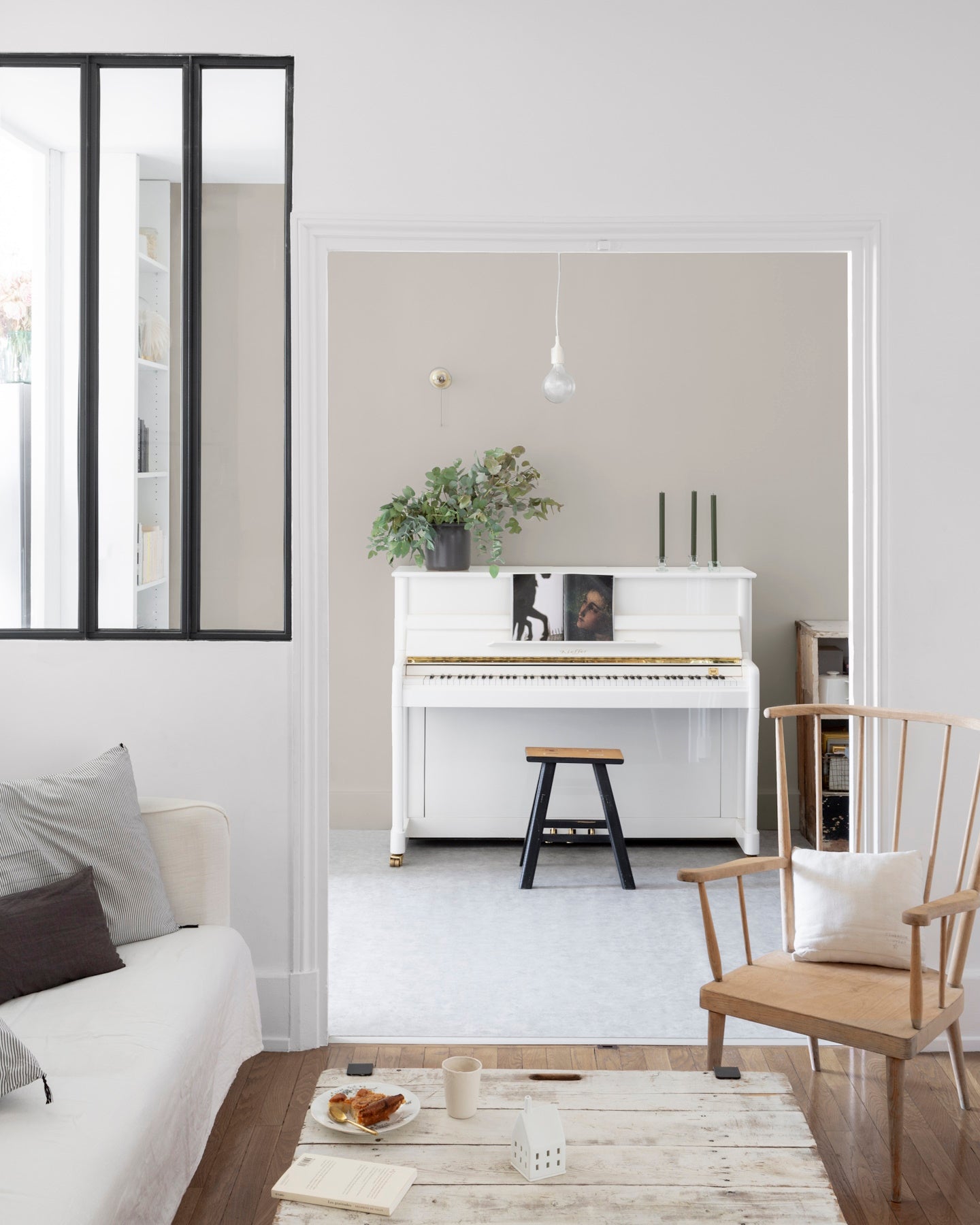 Salon aux murs blanc chiffon Libéron, avec verrière noire, piano blanc, canapé clair et chaise en bois.