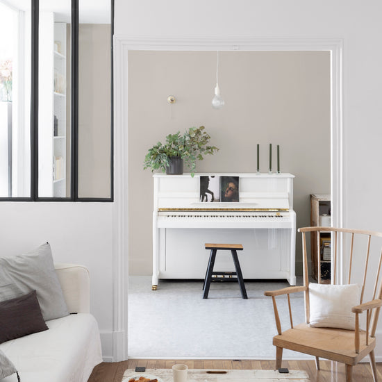 Salon aux murs blanc chiffon, Velours de Peinture Libéron, avec verrière noire, piano blanc et fauteuil en bois.