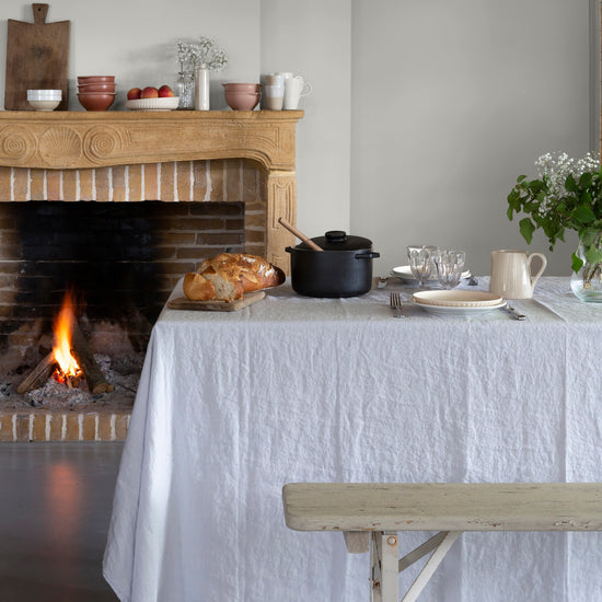 Salle à manger aux murs blanc grisaille, Velours de Peinture Libéron, avec cheminée en bois, table nappée et feu allumé.