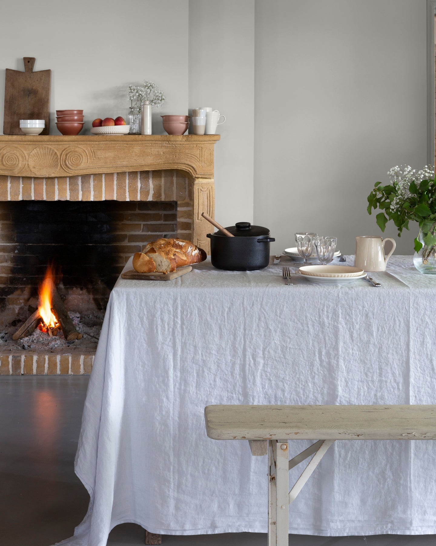Salle à manger aux murs blanc grisaille, Velours de Peinture Libéron, avec cheminée en bois, table nappée et feu allumé.