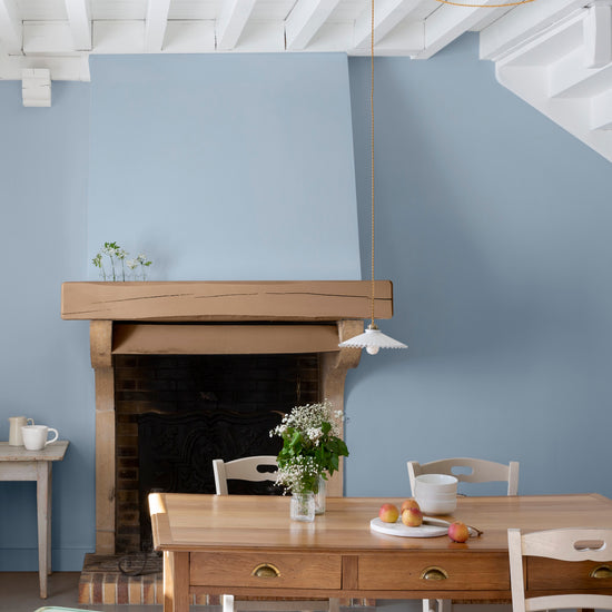 Salle à manger lumineuse aux murs peints en Bleu de Guède Velours de Peinture Libéron, avec cheminée en pierre, table en bois et mobilier aux tons naturels.