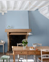 Salle à manger lumineuse aux murs peints en Bleu de Guède Velours de Peinture Libéron, avec cheminée en pierre, table en bois et mobilier aux tons naturels.