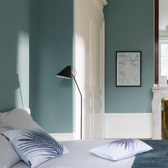 Chambre aux murs bleu Neptune Libéron, avec lit clair, lampadaire noir, porte blanche et cheminée.