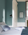 Chambre aux murs bleu Neptune Libéron, avec lit clair, lampadaire noir, porte blanche et cheminée.