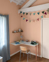 Chambre aux murs grès rosé, Velours de Peinture Libéron, avec bureau blanc, chaise en bois et guirlande colorée.