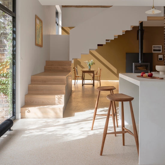 Pièce lumineuse aux murs gris saunier et bronze astrolabe Libéron, avec escalier en bois, cuisine blanche et tabourets.
