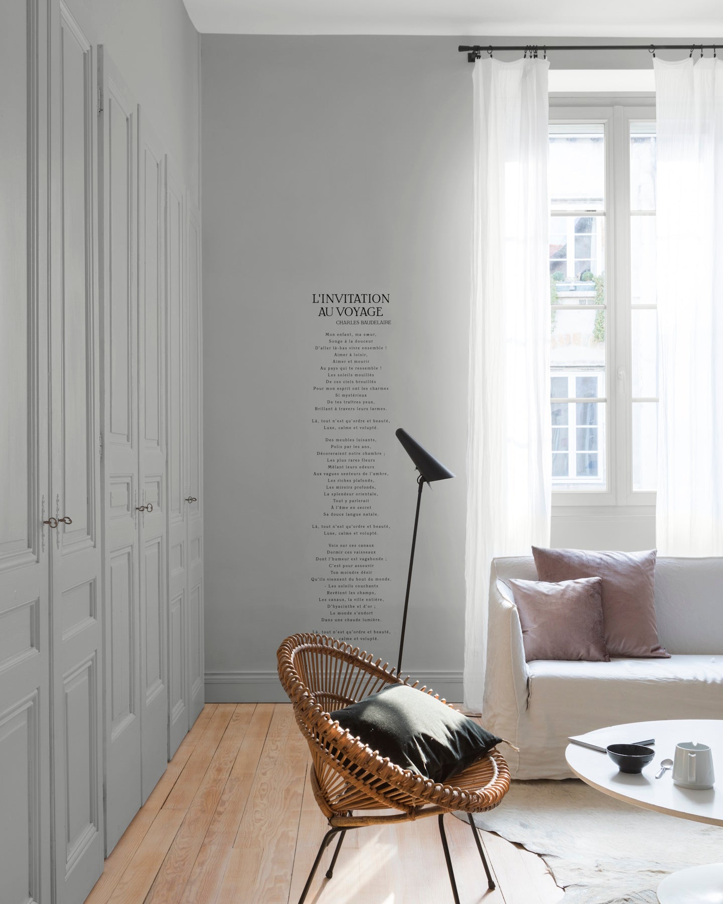Salon lumineux avec murs et portes de placards gris, peinture Velours de Peinture Gris Temps de Pluie Libéron, avec rideaux blancs, canapé gris et fauteuil en rotin