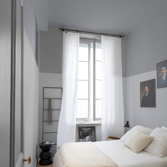 Chambre aux murs lumière opale et gris Gustave Libéron, avec lit blanc, rideaux légers et portraits encadrés.