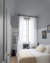 Chambre aux murs lumière opale et gris Gustave Libéron, avec lit blanc, rideaux légers et portraits encadrés.