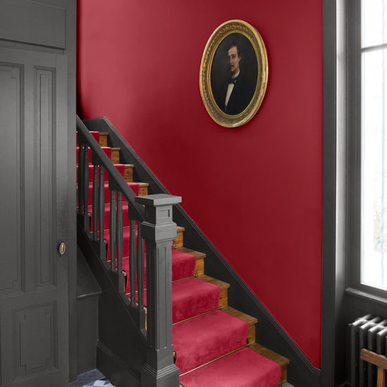 Cage d’escalier aux murs rouge Odéon et gris gravure Libéron, avec portrait ovale, escalier noir et tapis rouge.