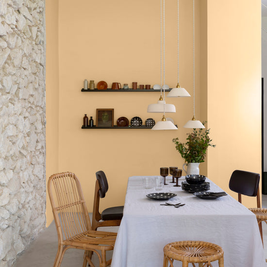 Salle à manger aux murs vanille bourbon, Velours de Peinture Libéron, avec chaises en rotin, table blanche et pierre apparente.