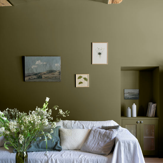 Salon aux murs vert bocage Libéron, avec canapé blanc, niche peinte, table en bois brut et bouquet champêtre.