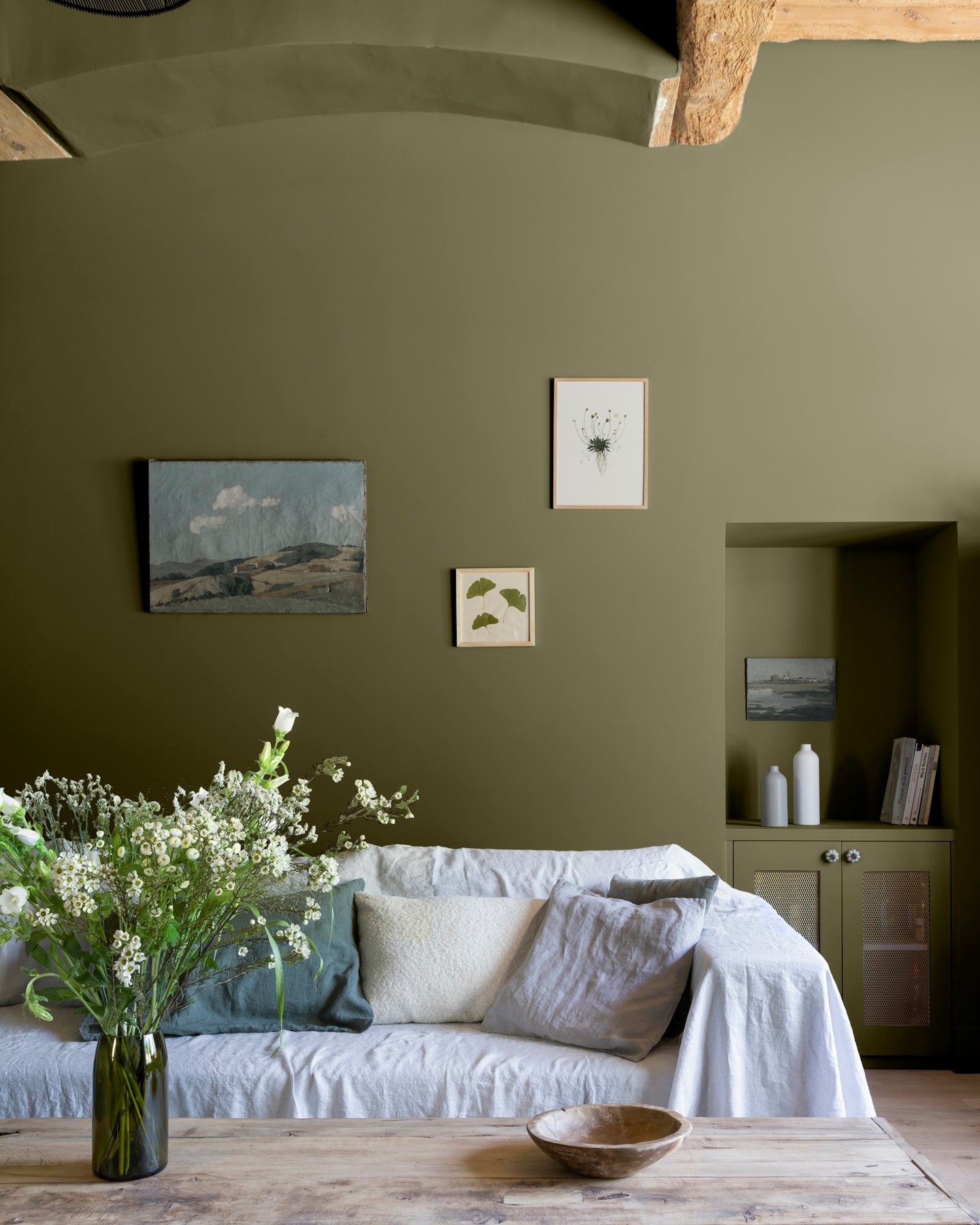 Salon aux murs vert bocage Libéron, avec canapé blanc, niche peinte, table en bois brut et bouquet champêtre.