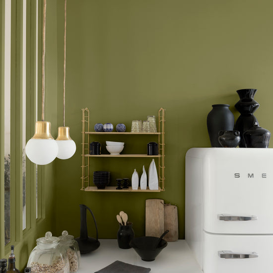 Cuisine aux murs vert herbarium, Velours de Peinture Libéron, avec étagère dorée, réfrigérateur blanc et suspensions.