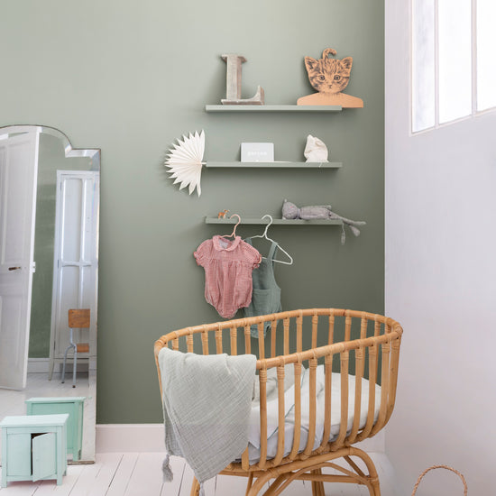 Chambre de bébé aux murs vert sauge Libéron, avec berceau en rotin, étagères murales, miroir et sol blanc.
