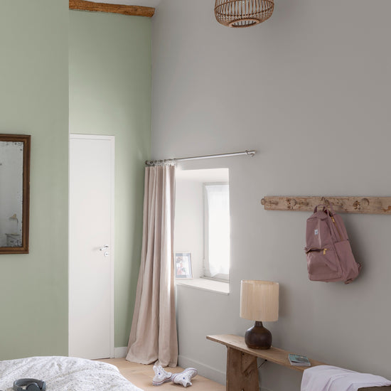 Chambre aux murs vert sauge et crème topinambour Libéron, avec banc en bois, rideaux poudrés et sac suspendu.