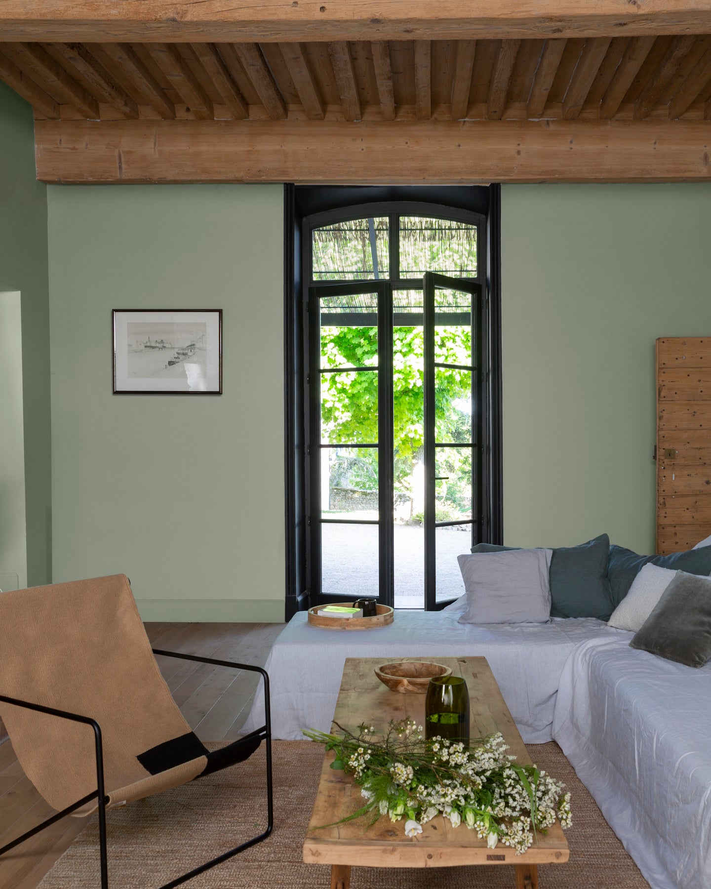 Salon aux murs vert sauge et noir smoking Libéron, avec poutres en bois, canapé blanc et baie vitrée noire.