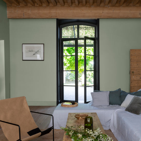 Salon aux murs vert sauge et fenêtre noir smoking Libéron, avec canapé blanc, fauteuil camel, table en bois et poutres apparentes.