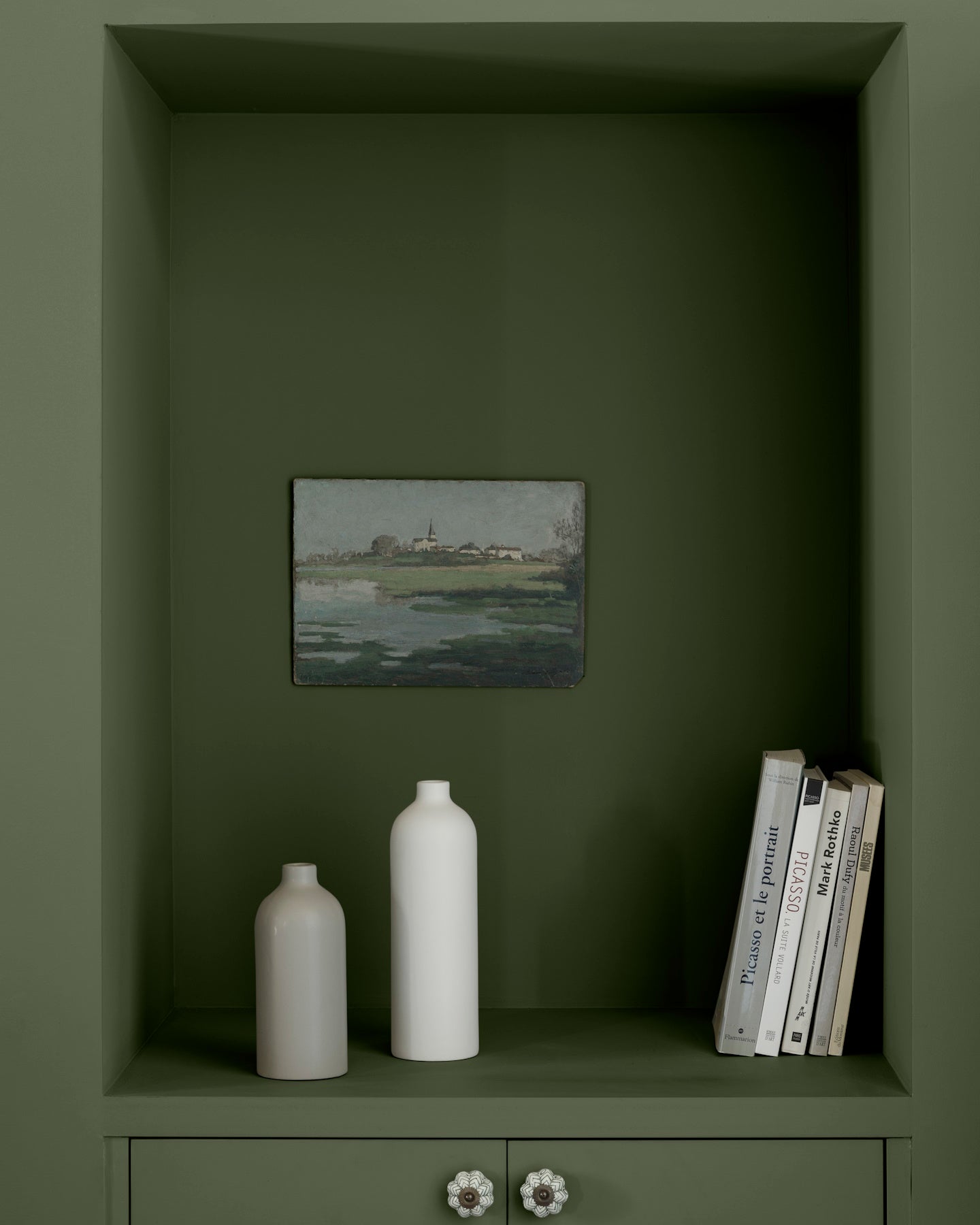 Niche murale vert treillis Libéron avec tableau de paysage, vases blancs et livres posés sur une étagère peinte.