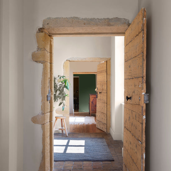 Couloir aux murs vert treillis et crème topinambour Libéron, avec portes en bois, pierre apparente et tapis rayé.